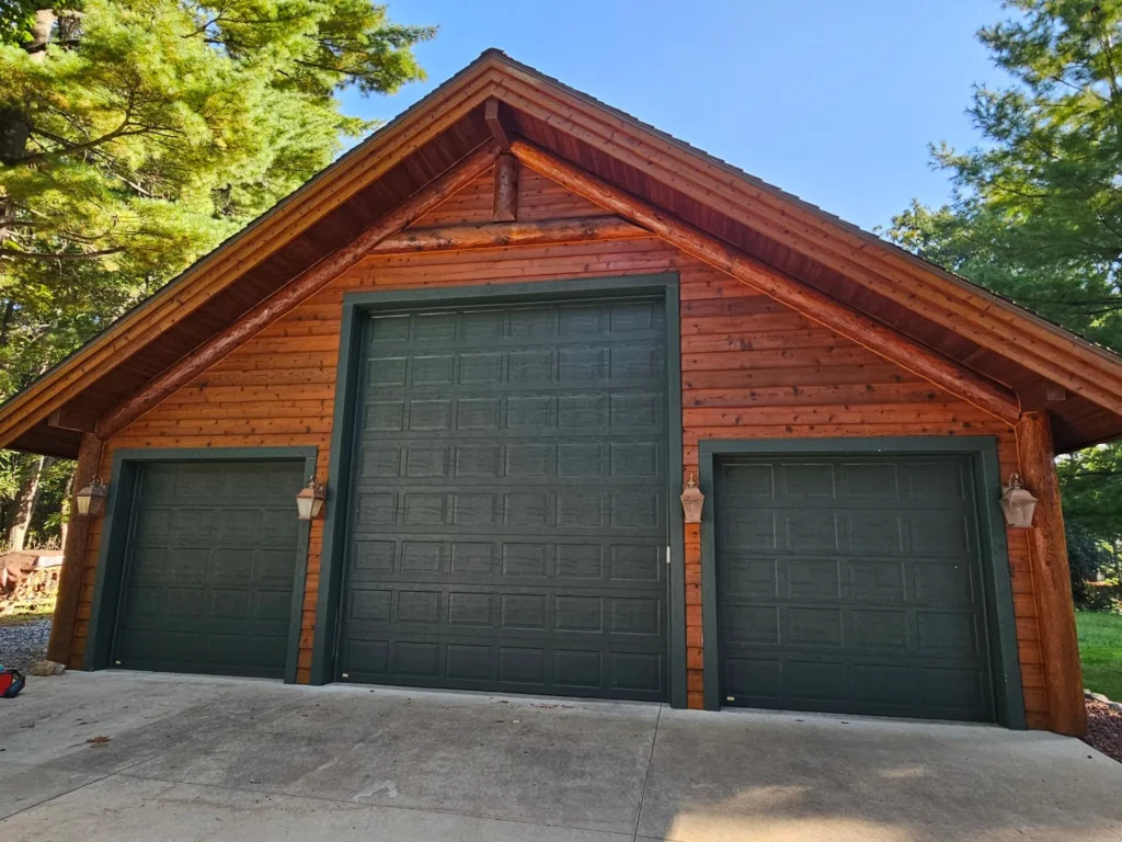 A wooden garage with three green doors is surrounded by tall trees, under a clear blue sky, with ample concrete driveway space.