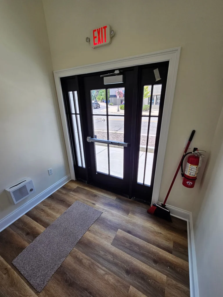 Interior scene showing an exit door with a window, a doormat, fire extinguisher, broom, and street view outside.