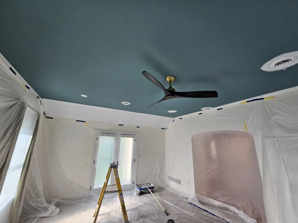 A room under renovation with plastic sheet coverings, blue ceiling, ceiling fan, ladder, and paint supplies near covered French doors.