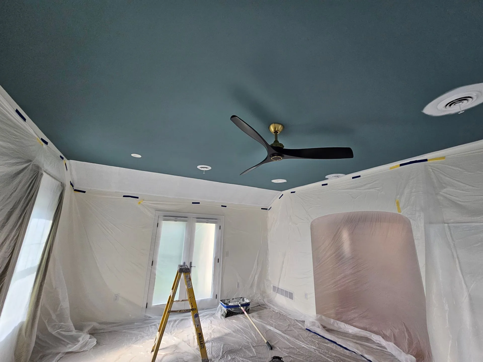 A room under renovation with plastic sheet coverings, blue ceiling, ceiling fan, ladder, and paint supplies near covered French doors.