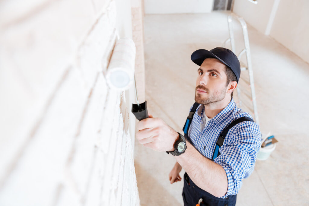 A person in a checkered shirt and cap paints a white wall with a roller in a spacious, unfinished room with a ladder.