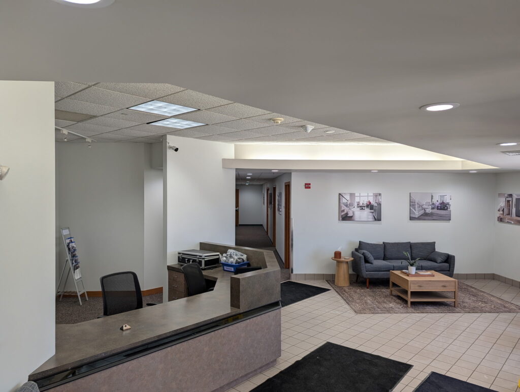 Modern office lobby with reception desk, waiting area with gray sofa, photographs on walls, and hallway leading to offices. Bright lighting overhead.
