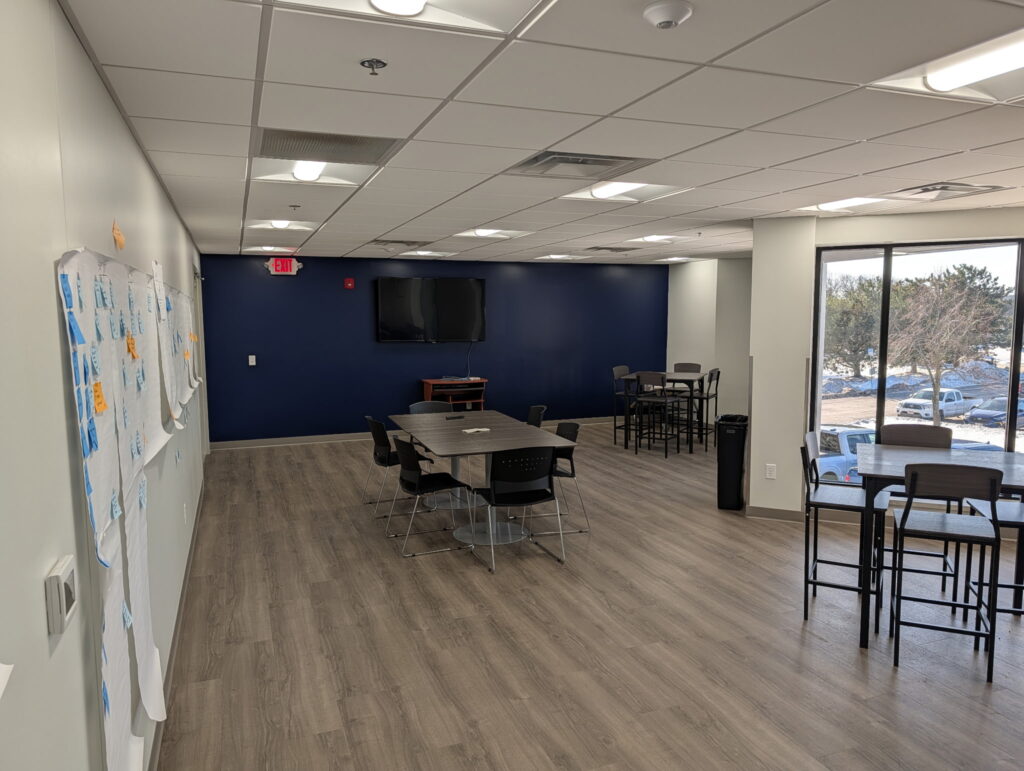 A modern office break room with tables, chairs, whiteboards, and a TV. Large windows overlook a parking lot with trees beyond.