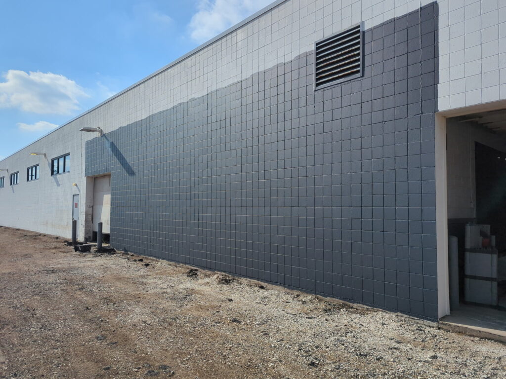 A long, partially painted building with black and white brickwork, exterior lamps, and open warehouse entrance on a gravel ground under blue sky.