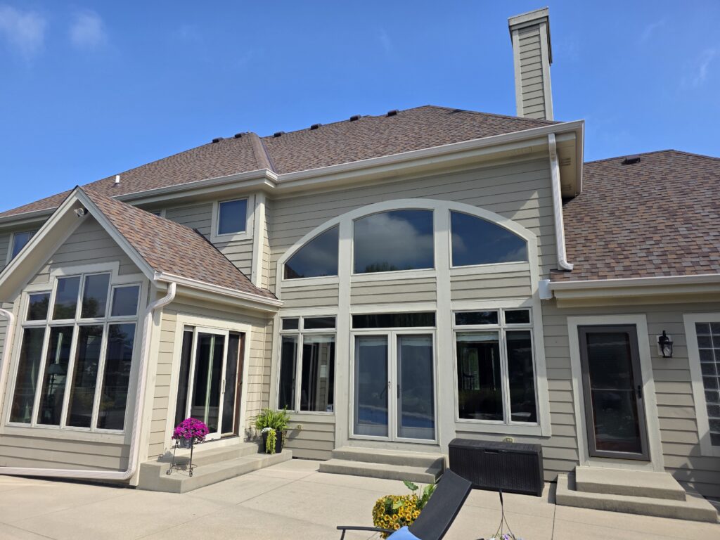 Modern two-story house with large windows, beige siding, and brown shingle roof. Bright patio features potted plants and black outdoor furniture.